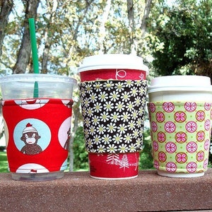 May include: Three reusable coffee cup sleeves with different patterns. The sleeve on the left has a red background with white circles and a cartoon monkey wearing a hat. The middle sleeve has a brown and white background with a daisy pattern. The sleeve on the right has a green background with pink and yellow circles.