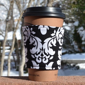 Könnte beinhalten: Eine Kaffeetassen-Hülle mit schwarzem und weißem Damastmuster. Die Hülle ist aus Stoff und hat einen weißen Hintergrund mit schwarzem Damastmuster. Die Hülle befindet sich auf einer braunen Kaffeetasse.