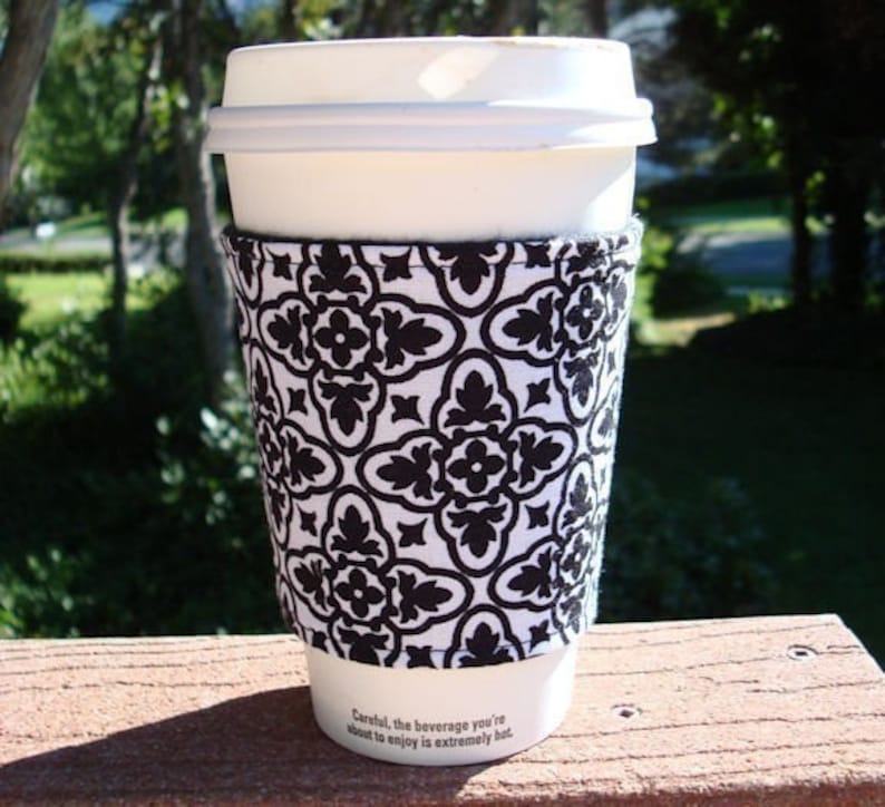 May include: A white to-go coffee cup with a black and white patterned fabric sleeve. The sleeve has a geometric design with a repeating pattern of black flowers and leaves on a white background. The text on the cup reads "Careful, the beverage you're about to enjoy is extremely hot."