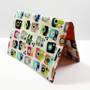 May include: A white fabric wallet with a colorful camera print. The wallet is folded in half and has a bright orange lining.