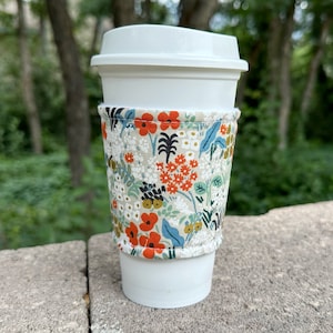 May include: A white to-go coffee cup with a floral print fabric sleeve. The sleeve has a beige background with orange, blue, and green flowers.