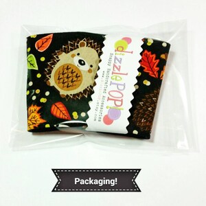 May include: A black and green fabric cup cozy with a hedgehog and autumn leaves design. The cozy is packaged in a clear plastic bag with a white label that reads "Dizzle Pop! Snappy Handcrafted Accessories www.dizzlepop.etsy.com".