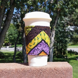 Könnte beinhalten: Ein weißer, wiederverwendbarer Kaffeebecher mit einer Stoffmanschette im Zickzackmuster in Schwarz, Limettengrün, Gelb, Violett und Orange. Der Stoff zeigt komplexe Linienmuster mit Schnörkeln und floralen Motiven. Der Becher hat einen weißen Deckel.