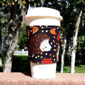 May include: A black and white coffee cup cozy with a hedgehog and leaf pattern. The cozy is made of fabric and has a white polka dot pattern. The text "Reusable, Recyclable, Re-enjoyable" is printed on the bottom of the cozy.