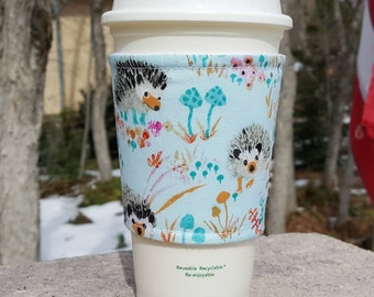 May include: A white reusable coffee cup with a blue and white fabric sleeve featuring a hedgehog and mushroom pattern. The sleeve has the text "Reusable. Recyclable. Re-enjoyable."
