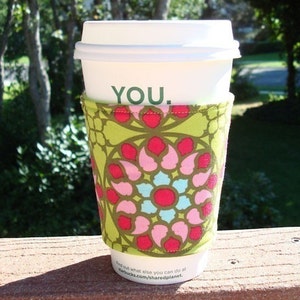 May include: A white coffee cup with a green and pink floral patterned sleeve. The cup has the word "YOU" printed on it.