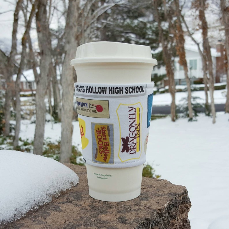 May include: A white to-go cup with a Stars Hollow High School themed reusable cup sleeve. The sleeve features a variety of designs, including the Dragonfly Inn, The Old Mill, and Stars Hollow Books.
