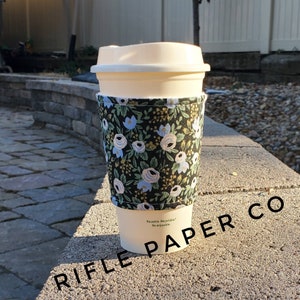May include: A white to-go coffee cup with a black and white floral fabric sleeve. The sleeve has a gold accent and the text "Rifle Paper Co." is printed on the bottom of the cup.