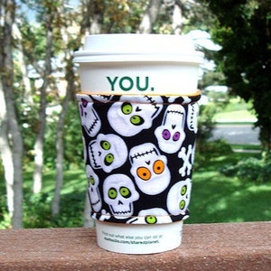 May include: A white disposable coffee cup with a white lid featuring the text YOU. in green. The cup is wrapped in a fabric sleeve with a black background and a repeating pattern of white skulls with green, orange, and purple eye sockets.