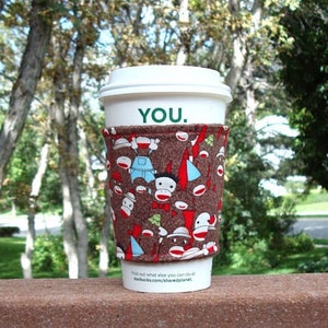May include: A white coffee cup with a brown and red fabric sleeve. The sleeve has a pattern of monkeys and the text "YOU." The cup is sitting on a wooden surface.