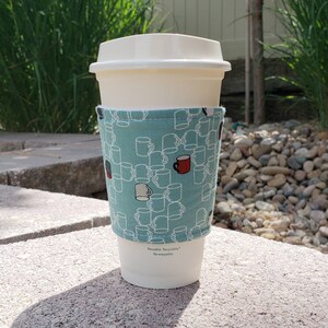 May include: A white reusable coffee cup with a teal fabric sleeve featuring a pattern of white and red mugs. The sleeve has the text "Reusable. Recyclable. Re-enjoyable." printed on the bottom.