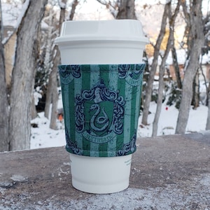 Könnte beinhalten: Eine grüne und blaue Kaffeetassenhülle mit einem Hogwarts-Wappen für Slytherin. Das Wappen zeigt eine Schlange und die Worte "Slytherin" auf einem Banner.