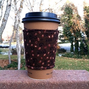 May include: A brown coffee cup with a black lid and a brown and pink coffee bean patterned sleeve.