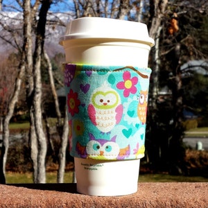 May include: A white reusable coffee cup with a colourful fabric sleeve featuring owls, flowers, and hearts. The sleeve has the text "Reusable, Recyclable, Re-enjoyable."