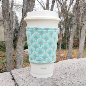 May include: A white to-go coffee cup with a teal and pink patterned fabric sleeve. The sleeve has a repeating pattern of teal fans with pink accents. The text on the cup reads "Reusable. Recyclable. Re-enjoyable."