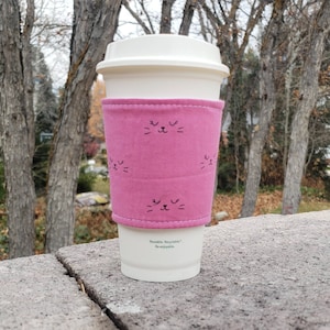 Puede incluir: Funda de taza de café reutilizable de color rosa con un diseño de cara de gato negro. La funda está hecha de tela y tiene una taza blanca en su interior.