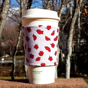 Pode incluir: Caneca de café branca com uma manga de tecido de padrão vermelho e branco. A manga tem um padrão repetido de lábios vermelhos e folhas. O texto na manga diz "Reutilizável. Reciclável. Reutilizável."