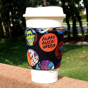 May include: A white reusable coffee cup featuring a black fabric sleeve with a Halloween print. The sleeve displays circular illustrations of a green monster, a skeleton, and a purple creature, with an orange circle reading HAPPY HALLOWEEN.