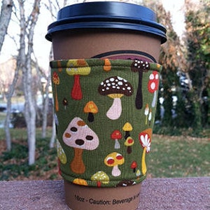 May include: A brown paper coffee cup with a black lid is wrapped with a green fabric sleeve. The sleeve is patterned with colorful mushrooms in various sizes and shapes. The cup is labeled "16oz - Caution: Beverage may be hot."