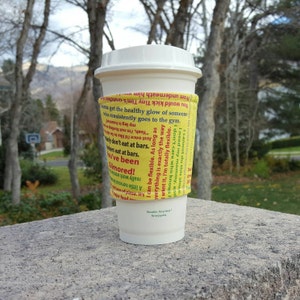 Może przedstawiać: Biały papierowy kubek do kawy z żółtym rękawem z tekstem. Rękaw ma cytaty i frazy w różnych kolorach. Kubek stoi na kamiennej powierzchni z rozmytym tłem zewnętrznym.