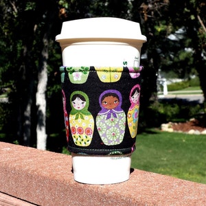 Op de afbeelding: Een witte to-go koffiebeker met een zwarte en kleurrijke stoffen hoes met een patroon van Russische poppen. De hoes heeft een zwarte achtergrond met kleurrijke Russische poppen in verschillende tinten roze, groen, geel en blauw. De tekst "Dit product is mogelijk niet vaatwasmachinebestendig" staat gedrukt op de bodem van de beker.