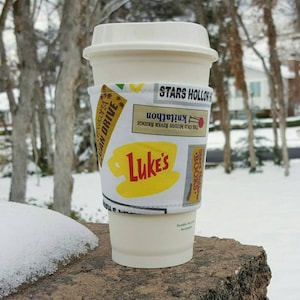 Può includere: Una tazza di caffè bianca con un manicotto in tessuto a tema Stars Hollow. Il manicotto presenta le parole "Stars Hollow", "The Old Muddy River Bridge", "Knitathon" e "Luke's".