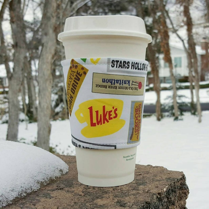 May include: A white coffee cup with a Stars Hollow themed fabric sleeve. The sleeve features the words "Stars Hollow", "The Old Muddy River Bridge", "Knitathon", and "Luke's".