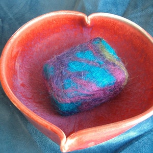 May include: A red ceramic bowl with a heart-shaped rim, containing a colourful wool soap. The soap is a mix of purple, blue, and green.