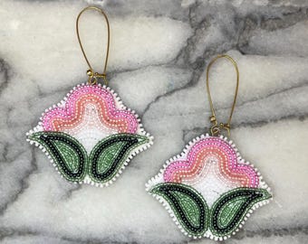 Op de afbeelding: Een paar kralen oorbellen in een bloemenontwerp. De oorbellen hebben een roze en witte bloemvorm met groene bladaccenten. De oorbellen hebben goudkleurige oorhaken en zijn geplaatst tegen een marmeren achtergrond.