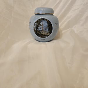 Op de afbeelding: Een lichtblauwe keramische pot met deksel, met een gedetailleerde illustratie van een kathedraal. De pot heeft een gouden rand en staat tegen een witte achtergrond.