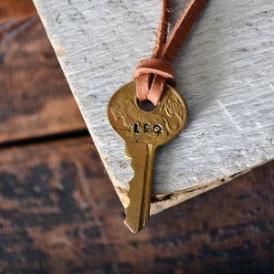 May include: A brass key with the word "LEO" engraved on it is attached to a brown leather cord. The key is hanging on a white wooden surface.