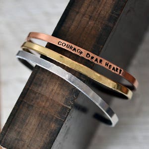 Könnte beinhalten: Drei übereinanderliegende Metallarmbänder mit gehämmerter Oberfläche. Das obere Armband ist aus Kupfer und trägt die eingravierten Worte "Courage Dear Heart".