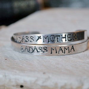 May include: Two silver bracelets stacked on top of each other. The top bracelet says "Badass Mother" and the bottom bracelet says "Badass Mama".