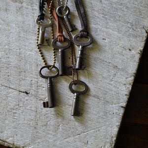 May include: A set of six antique keys on leather cords and chains. The keys are silver and have a worn, rustic look. The cords are brown and black.