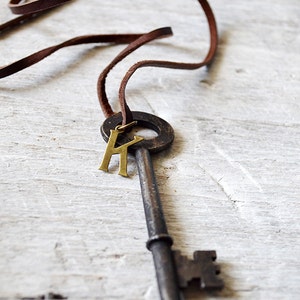 May include: A brown leather cord necklace with a large antique-style key pendant. The key has a brass letter "K" charm attached to the ring.
