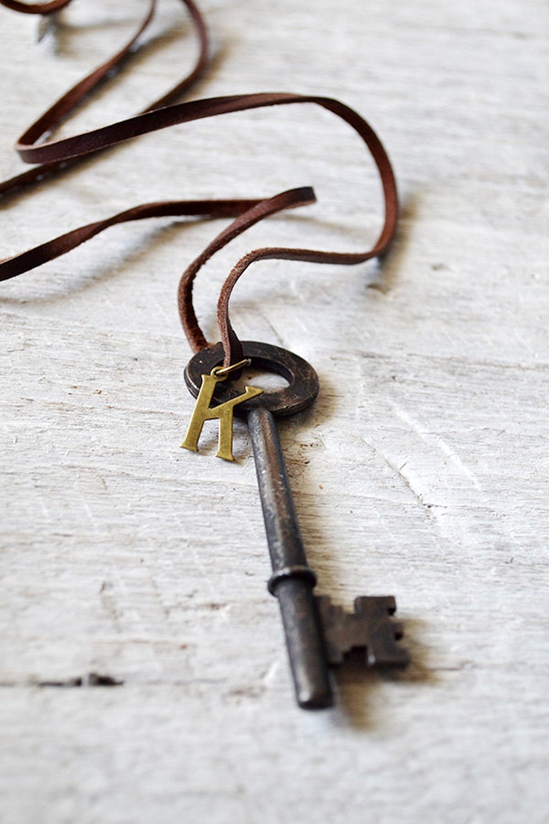 May include: A brown leather cord necklace with a large antique-style key pendant. The key has a brass letter "K" charm attached to the ring.