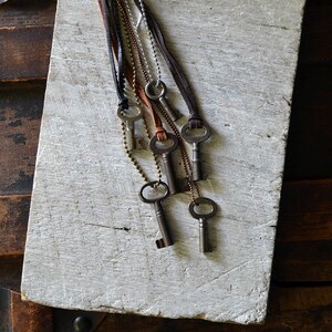 May include: A set of six antique keys on leather and chain necklaces. The keys are silver and have different shapes and sizes. The necklaces are hanging on a white wooden surface.