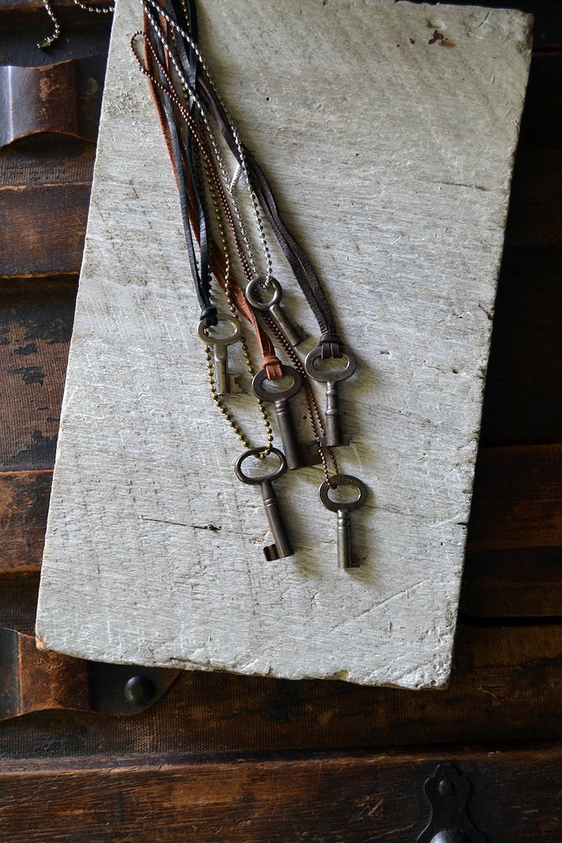 May include: A set of six antique keys on leather and chain necklaces. The keys are silver and have different shapes and sizes. The necklaces are hanging on a white wooden surface.