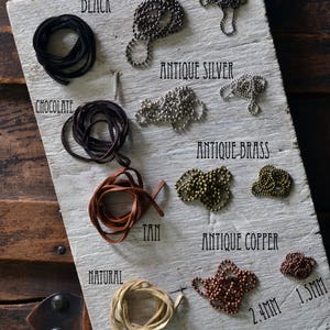 May include: A white wooden board with different colors of leather cord and metal chain. The colors include black, chocolate, tan, natural, gunmetal, antique silver, antique brass, and antique copper. The chains are labeled with their sizes in millimeters. The board is labeled with the names of the colors and materials.