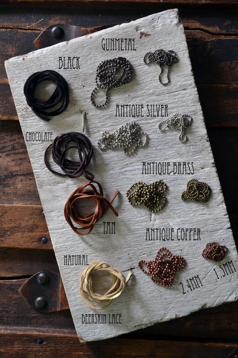 May include: A white wooden board with different colors of leather cord and metal chain. The colors include black, chocolate, tan, natural, gunmetal, antique silver, antique brass, and antique copper. The chains are labeled with their sizes in millimeters. The board is labeled with the names of the colors and materials.