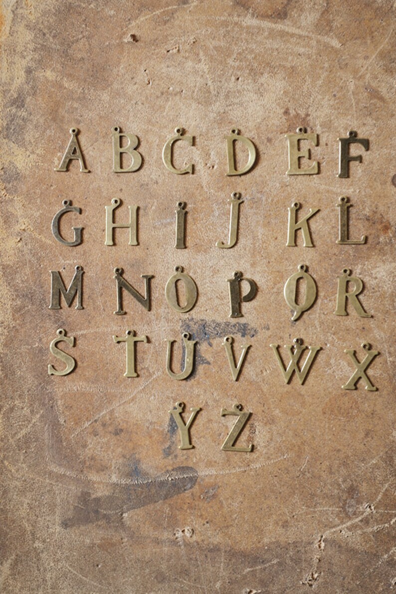 May include: A set of 26 brass letter charms, each letter of the alphabet is displayed in a row on a brown surface. The letters are all the same size and style, and they have a slightly aged look.