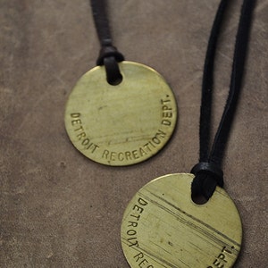 May include: Two gold-colored round pendants with black leather cords. The pendants are engraved with the text "DETROIT RECREATION DEPT."