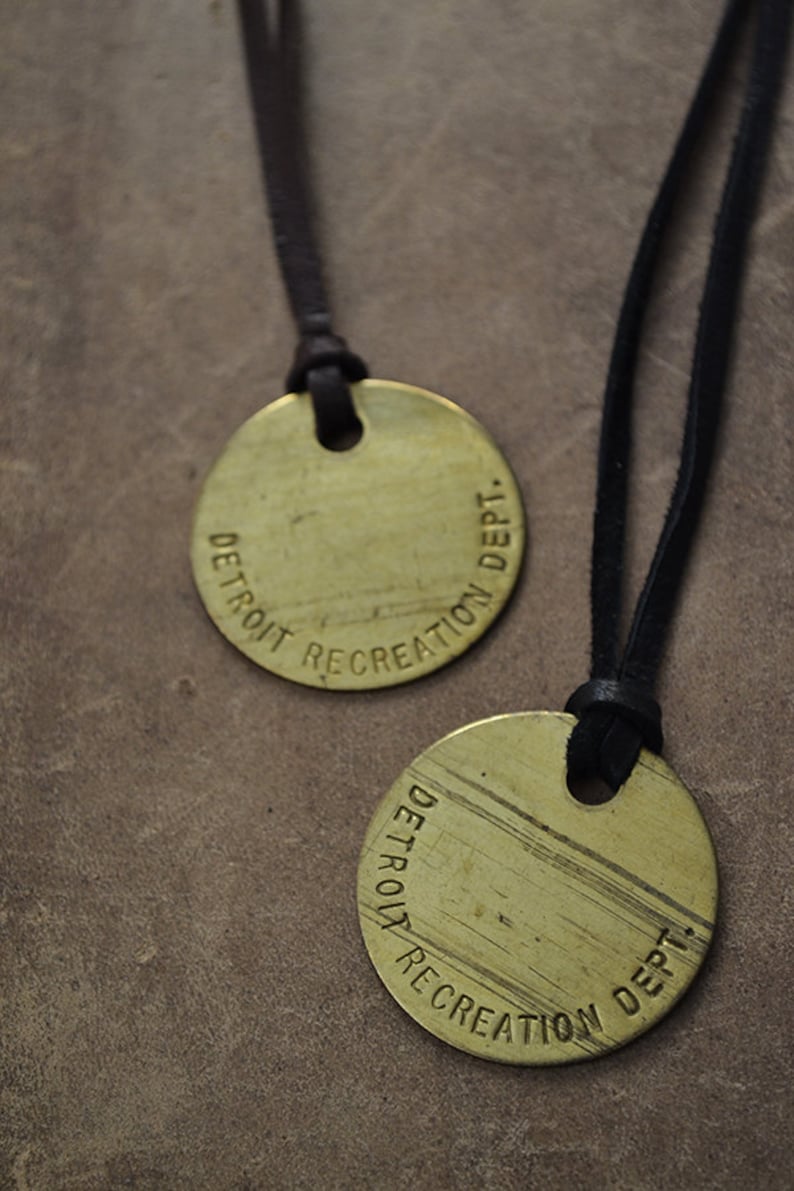 May include: Two gold-colored round pendants with black leather cords. The pendants are engraved with the text "DETROIT RECREATION DEPT."