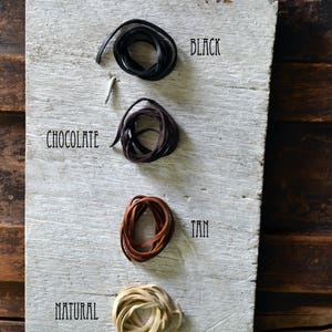 May include: Four spools of leather cord in different colors: black, chocolate brown, tan, and natural. The cords are arranged vertically on a white wooden surface.