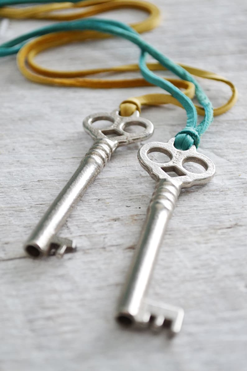 Op de afbeelding: Twee zilveren skeletsleutels aan een turquoise en gele koord. De sleutels hebben ingewikkelde ontwerpen en zijn met een knoop aan elkaar gebonden.