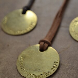 May include: A brass pendant with a brown leather cord. The pendant is round and has the text "DETROIT RECREATION DEPT." engraved on it.