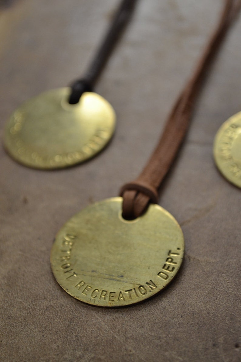 May include: A brass pendant with a brown leather cord. The pendant is round and has the text "DETROIT RECREATION DEPT." engraved on it.