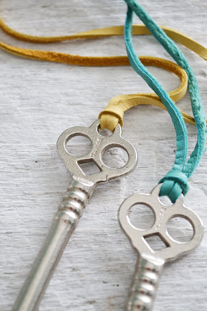 Op de afbeelding: Twee zilveren skeletsleutels met sierlijke sleutelgaten en een decoratieve, gedraaide steel. De sleutels zijn bevestigd aan een teal- en gele leren koord.
