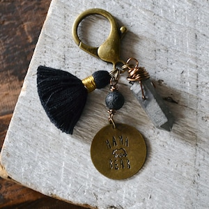 May include: A brass keychain with a black tassel, a lava rock bead, a grey crystal, and a brass charm that says "Mama Bear" with a bear silhouette.