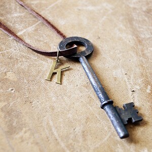 May include: A brown leather cord necklace with a large antique-style key pendant and a gold letter 'K' charm.